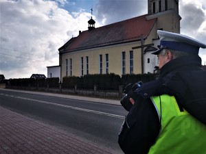 policjant drogówki w odblaskowej kamizelce stoi przy drodze i dokonuje pomiaru prędkości pojazdów. W tle policyjny orzeski kościół