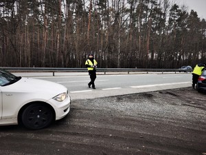 policjant drogówki w odblaskowej kamizelce stoi przy drodze i zatrzymuje do kontroli biały samochód osobowy