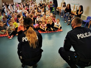 policjanci (kobieta i mężczyzna) w trakcie zajęć profilaktycznych z przedszkolakami. Scena ma miejsce w jednej z sal. Dzieci siedzą na dywaniku