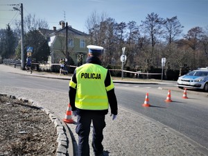 stojący tyłem, w odblaskowej kamizelce policjant w trakcie zabezpieczenia. Na drodze stoją pachołki