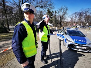 policjanci drogówki stoją przed radiowozem. Jeden z nich wskazuje na coś palcem