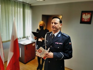 komendant Policji trzyma w dłoniach konstytucję, obok niego flagi Polski