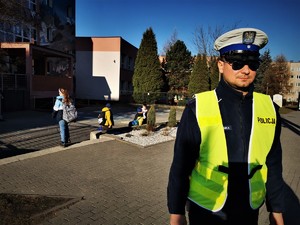 policjant ruchu drogowego stoi przed budynkiem szkoły, w tle dzieci, mundurowy ma założone okulary przeciwsłoneczne