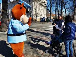 policyjna maskotka wraz z policjantką rozmawiają z przechodniami w centrum miasta, w tle przejeżdzające samochody