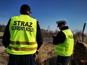 stojący przy drodze policjant oraz funkcjonariusz straży ochrony kolei. Obaj w odblaskowych kamizelkach