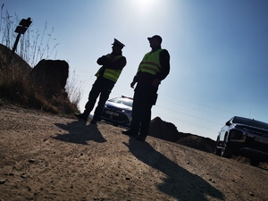 stojący przy drodze policjant oraz funkcjonariusz straży ochrony kolei. Obaj w odblaskowych kamizelkach. Świeci słońce