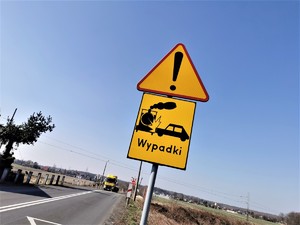 znak ostrzegawczy &quot;UWAGA WYPADKI Z UDZIAŁEM POCIĄGU&quot;, w tle przejazd kolejowy. Jest słoneczny dzień