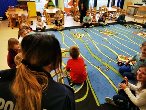 policjantka prowadzi zajęcia z dziećmi. Maluchy siedzą na dywaniku, zajęcia odbywają się w sali przedszkolnej
