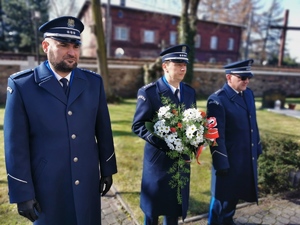 trzech policjantów w mundurach wyjściowych stoi przed tablicą upamiętniającą pomordowanych policjantów. Mikołów, dzielnica Bujaków. Policjant stojący w środku trzyma bukiet kwiatów