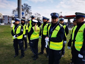 stojący w grupie policjanci, wszysycy mają założone odblaskowe kamizelki. W tle stacja paliw