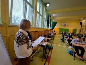 sala gimnastyczna, rozłożone ławki, w nich uczniowie, kobieta tłumaczy zasady rozwiązywania testu