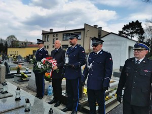 cmentarz parafialny w Łaziskach Górnych. Stojący w rzędzie policjanci w mundurach galowych oraz pozostali uczestnicy uroczystości. Policjant trzyma znicz, starosta trzyma kwiaty