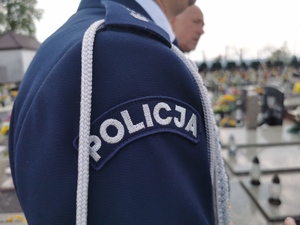 zbliżenie na policyjny mundur i na napis POLICJA, w tle groby