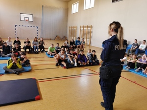 sala gimnastyczna w szkole, dzieci siedzące na matercach, które słuchają porad o bezpieczeństwie. W tle policjantka