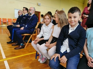 siedzące na ławkach małe dzieci, obok nich policjanci w mundurach wyjściowych