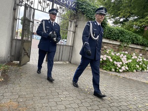 dwóch policjantów w mundurach galowych wchodzi na teren przy kościele, jeden z nich trzyma znicz