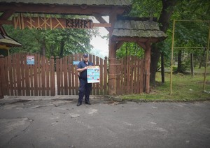 dzielnicowy przed wejściem na teren ośrodka &quot;Baraniok&quot;. Mężczyzna trzyma plakat promnujący kampanię &quot;Kręci mnie bezpieczeństwo nad wodą&quot;