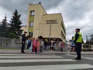 grupa dzieci stoi na chodniku przed oznakowanym przejściem dla pieszych, w tle budynek szkoły