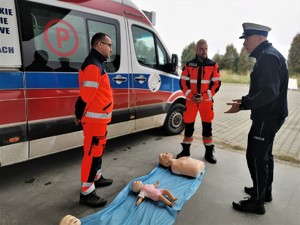 zdjęcie kolorowe: dwóch ratowników medycznych rozmawia z policjantem drogówki, w tle karetka pogotowia, na ziemi leżą fantomy