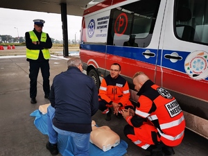 dwóch ratowników medycznych oraz policjant drogówki przygląda się wykonywaniu zadania z pierwszej pomocy zatrzymanemu do kontroli drogowej kierującemu