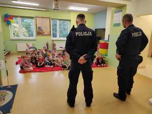 sala w przedszkolu. Siedzące na podłodze dzieci, przed nimi dwaj dzielnicowi