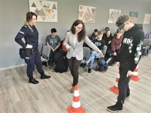 młodzież szkolna podczas zajęć praktycznych. Scena ma miejsce w budynku szkoły. Na środku sali rozstawione pachołki. Widać też policjantkę oraz pracownicę Sanepidu