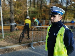 policjant drogówki w odblaskowej kamizelce, w tle uczestnicy wyścigu kolarskiego. Jest słoneczny dzień