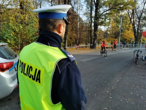 policjant drogówki w odblaskowej kamizelce, w tle uczestnicy wyścigu kolarskiego. Jest słoneczny dzień, godziny poranne