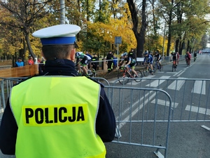 policjant drogówki w odblaskowej kamizelce, w tle uczestnicy wyścigu kolarskiego. Jest słoneczny dzień