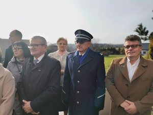 policjant stojący przy przedstawicielach samorządu w trakcie odśpiewania hymnu państwowego. Scena ma miejsce przed budynkiem kościoła