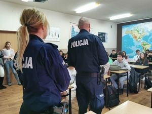 Na zdjęciu widać dwoje policjantów. Sylwetki od tyłu. W tle uczniowie