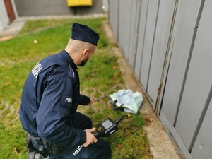 na zdjęciu reklamówka oraz policjant dokonujący sprawdzenia pakunku, widać też fragment blaszanego garażu