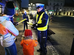 na zdjęciu widać policjanta drogówki, który rozdaje odblaskowe opaski przechodniom. Scena ma miejsce na płycie mikołowskiego rynku