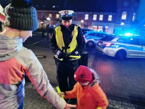 na zdjęciu widać policjanta rozmawiającego z kobietą i jej dziećmi. Mundurowy ma założoną odblaskową opaskę