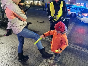 na zdjęciu widać małe dziecko, które zakłada mamie odblaskową opaskę na nogę. Jest ciemno.