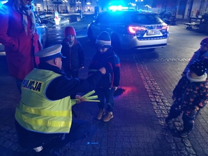 na zdjęciu widać policjanta w odblaskowej kamizelce, który daje odblaskową opaskę dziecku. Scena ma miejsce na płycie mikołowskiego rynku