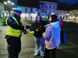 na zdjęciu widać policjanta drogówki, który przekazuje odblaskową opaskę dwóm nastolatkom