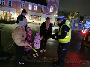 na zdjęciu widać policjanta drogówki, który przekazuje przechodniom odblaskowe opaski. Jest zmrok