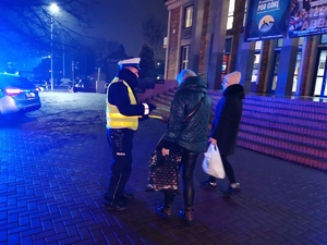 policjant drogówki rozdaje odblaskowe opaski przechodniom. Panuje zmrok.