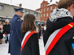 na zdjęciu widać policjanta w mundurze galowym oraz uczniów szkoły z przewieszonymi szarfami w kolorze biało-czerwonym