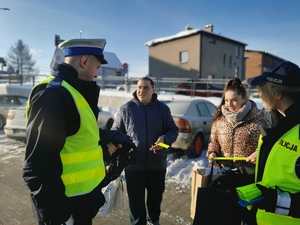 na zdjęciu widać dwoje policjantów, którzy rozdawają przechodniom odblaskowe opaski. Scena ma miejsce na parkingu w centrum Łazisk Górnych