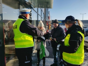 na zdjęciu widać dwoje policjantów przed sklepem, którzy wręczają przechodniom opaski