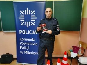 na zdjęciu policjant prewencji, który trzyma w ręce ulotki kampanii antynarkotykowej, tuż za nim baner koloru granatowego z napisem :Komenda Powiatowa Policji w Mikołowie