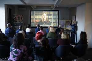 na zdjęciu jedna z sal bibliotecznych, na rzutniku wyświetlany jest film, przemawia nastolatka, uczestnicy konferencji siedzą na krzesłach i oglądają film