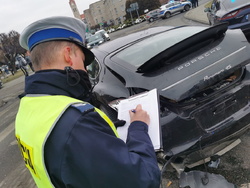 Na kolorowym zdjęciu widoczny policjant sporządzający dokumentację na miejscu zdarzenia. Za nim widoczny rozbity czarny samochód.