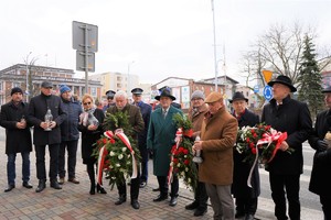 na zdjęciu kilkanaście osób stoi przez tablicą przy urzędzie. Niektóre osoby trzymają znicze, inne wiązanki kwiatów