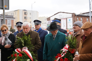 na zdjęciu kilkanaście osób stoi przez tablicą przy urzędzie. Niektóre osoby trzymają znicze, inne wiązanki kwiatów. Wśród nich są policjanci