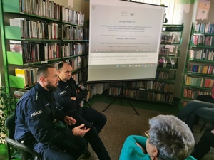 na zdjęciu widać salę biblioteczną, w której siedzą dwaj policjanci, wyświetlana jest prezentacja, po prawej stronie siedzą seniorzy. Scena ma miejsce w budynku Gminnej Biblioteki w Wyrach