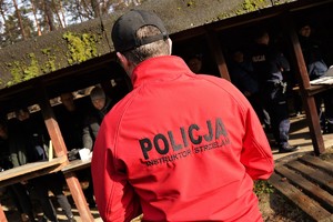 na zdjęciu widać instruktora wyszkolenia strzeleckiego. Mężczyzna ma na sobie polar koloru czerwonego z czarnymi napisami POLICJA