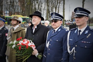 policjanci w mundurach galowych oraz pozostali mundurowi i przedstawiciele samorządu stoją  w rzędzie na cmentarzu w Ornontowicach. Starosta trzyma wiązankę kwiatów